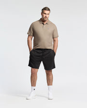 A man in Psycho Bunny's Men's Big and Tall Classic Pique Polo Shirt, style B9K001CRPC, and black shorts stands with hands in pockets against a plain backdrop. Sporting short hair, a beard, white socks, and black shoes completes his look.
