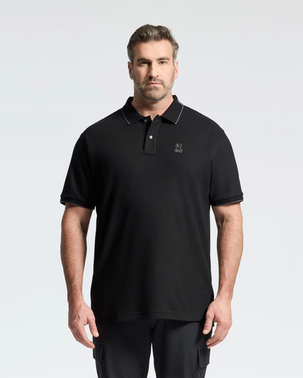 A bearded man with short hair stands against a plain background, wearing Psycho Bunny's Mens Big and Tall New Castle Pique Polo (B9K487F200), crafted from 100% Pima cotton. The luxury black shirt has an embroidered chest logo, his hands resting by his sides.