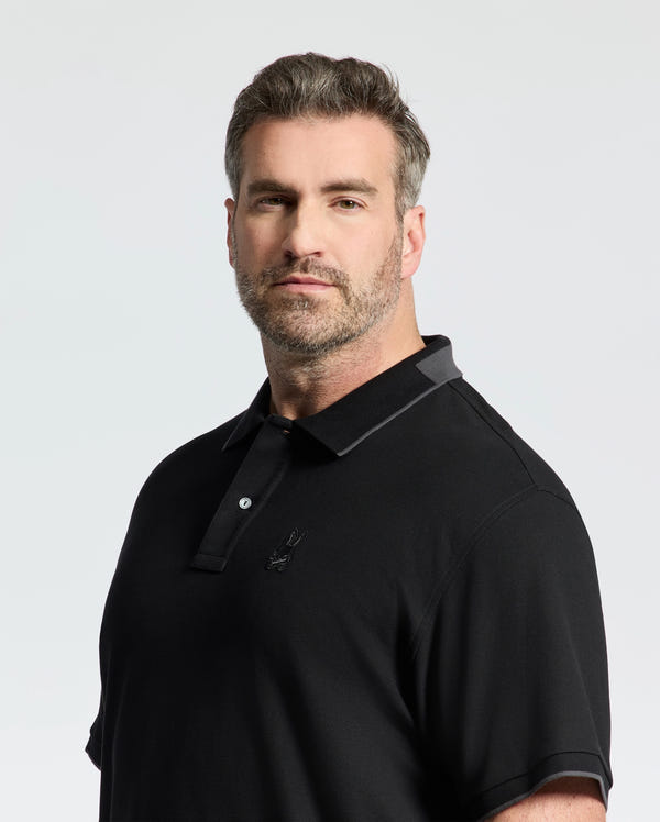 A man with short hair and a beard wears a Psycho Bunny MENS BIG AND TALL NEW CASTLE PIQUE POLO - B9K487F200, made from 100% Pima cotton with luxury finishes and an emblem on the chest. He stands against a plain light background, looking slightly to the side.
