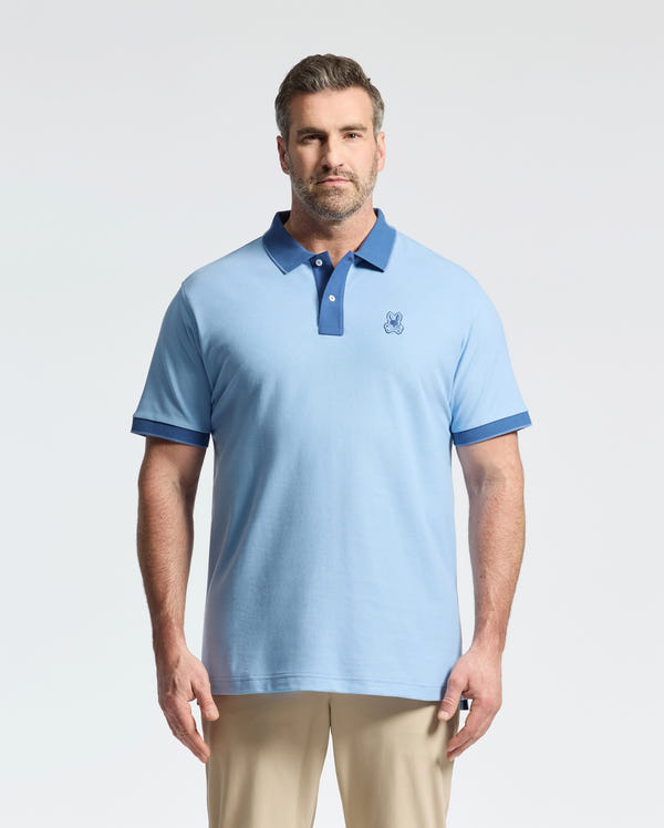A man stands facing forward in a slim fit light blue Psycho Bunny MENS BIG AND TALL NEW CASTLE PIQUE POLO - B9K487F200 with dark blue accents and beige pants, set against a plain light background.