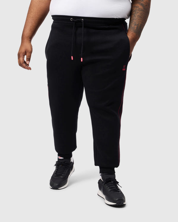 A person models Psycho Bunny's BIG AND TALL TRAVIS TRACK PANT - B9P143D200 in black with pink accents and an elastic waistband, paired with sleek black sneakers and a crisp white shirt. Set against a plain gray background, their arm tattoo adds intrigue to the relaxed yet stylish look.