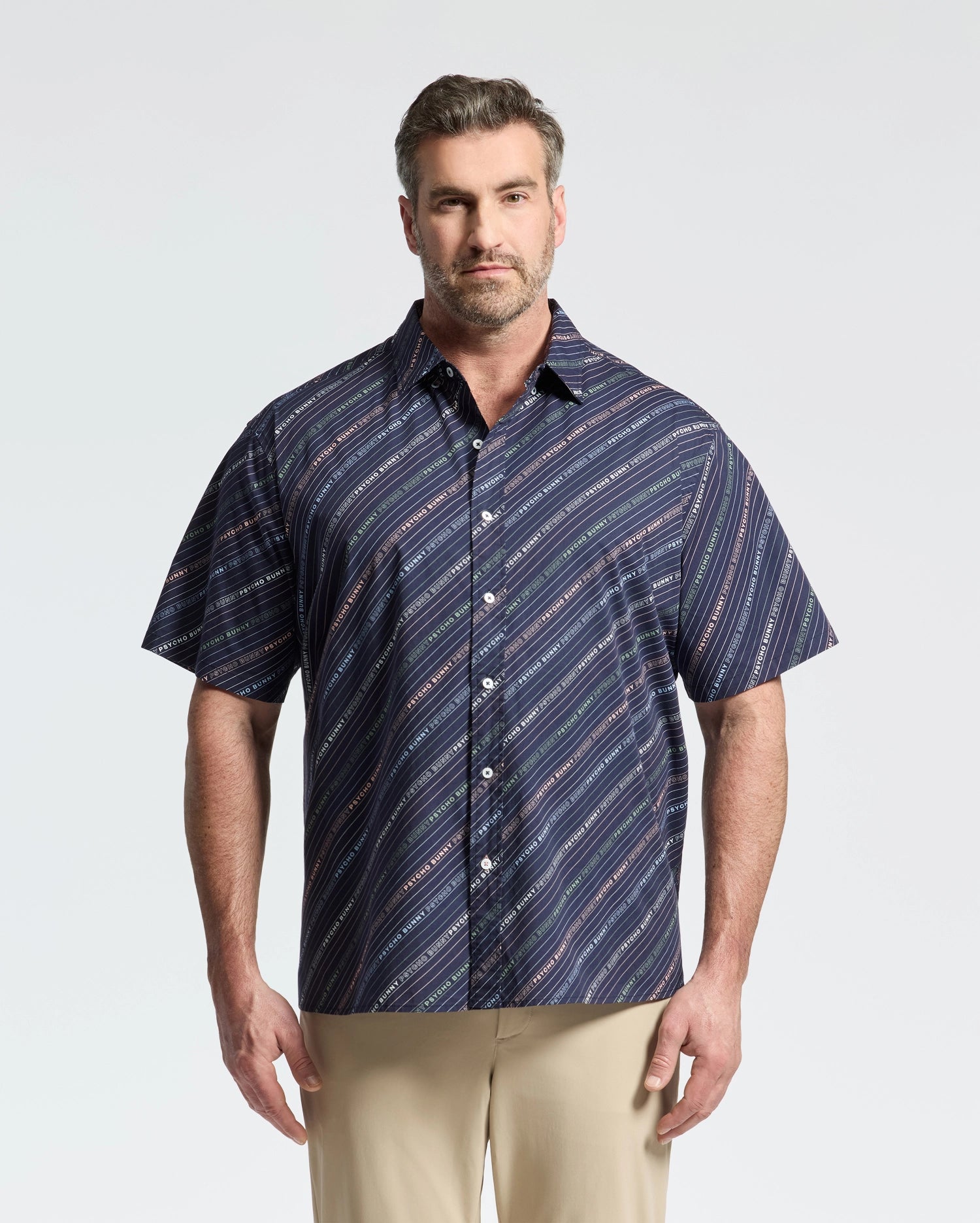A man sports a Psycho Bunny MENS BIG AND TALL RENMARK STRETCH POPLIN WOVEN SHIRT in blue diagonal stripes, stylishly paired with beige pants against a light backdrop.