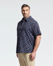 A man confidently poses in a Psycho Bunny Men's Big and Tall Renmark Stretch Poplin Woven Shirt (B9Q629F200), showcasing a navy blue and white diagonal stripe design, with neatly styled hair. He pairs it with light khaki pants against a plain background.
