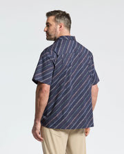 A man, sporting short hair and a beard, stands sideways wearing Psycho Bunny's MENS BIG AND TALL RENMARK STRETCH POPLIN WOVEN SHIRT - B9Q629F200, featuring a navy diagonal striped design. He pairs it with light-colored pants against a plain white background.