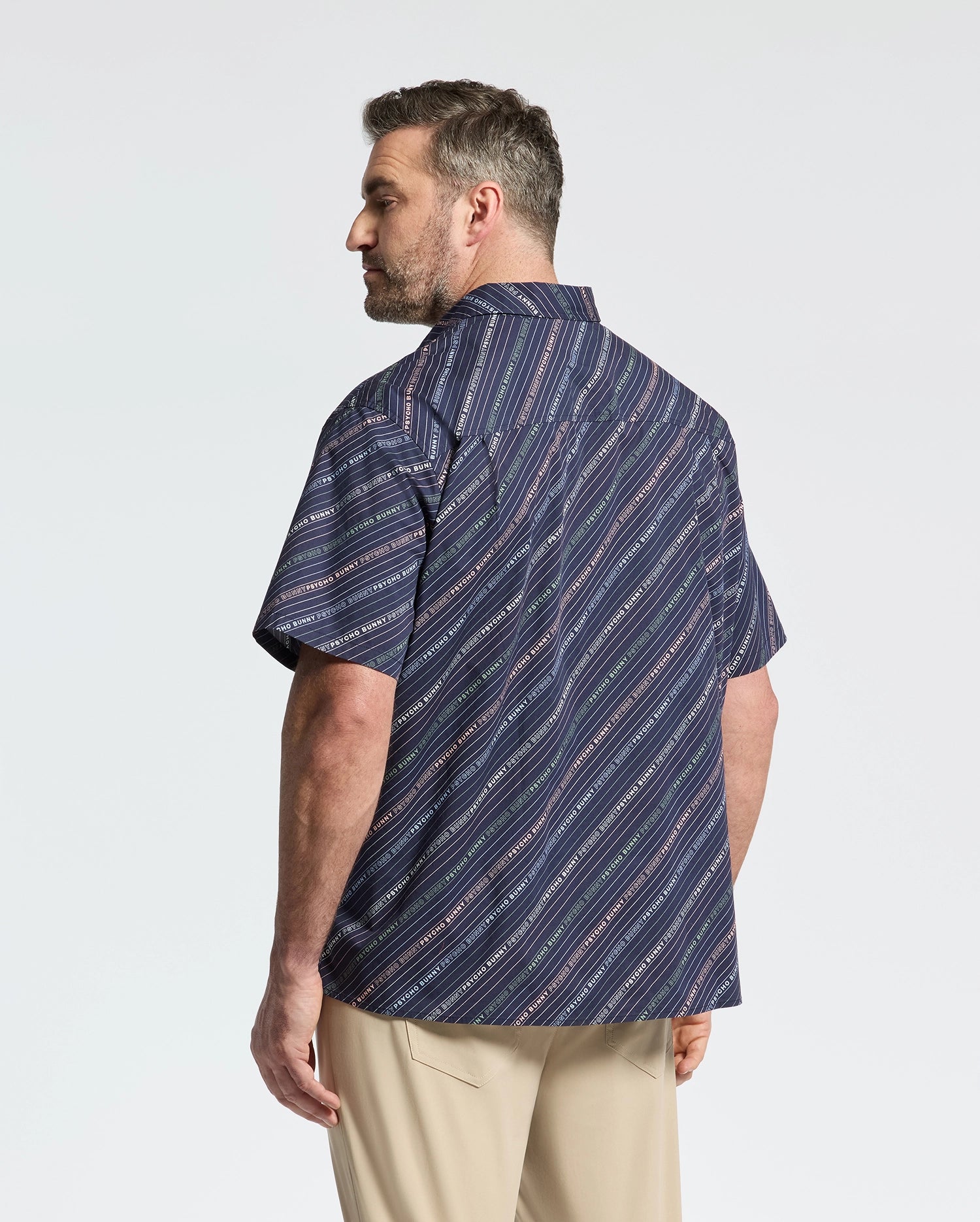 A man, sporting short hair and a beard, stands sideways wearing Psycho Bunny's MENS BIG AND TALL RENMARK STRETCH POPLIN WOVEN SHIRT - B9Q629F200, featuring a navy diagonal striped design. He pairs it with light-colored pants against a plain white background.