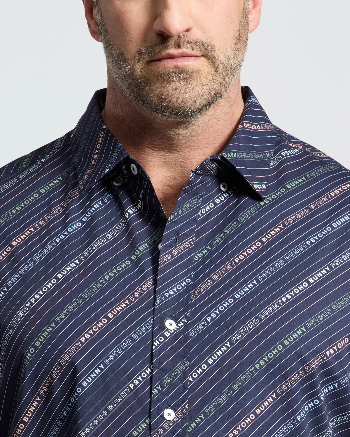 A bearded man wears a Psycho Bunny Renmark short sleeve shirt, featuring a logo-patterned print in vibrant colors against dark blue diagonal stripes. The premium stretch poplin collared shirt contrasts with the plain light gray backdrop.