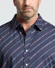 A bearded man wears a Psycho Bunny Renmark short sleeve shirt, featuring a logo-patterned print in vibrant colors against dark blue diagonal stripes. The premium stretch poplin collared shirt contrasts with the plain light gray backdrop.