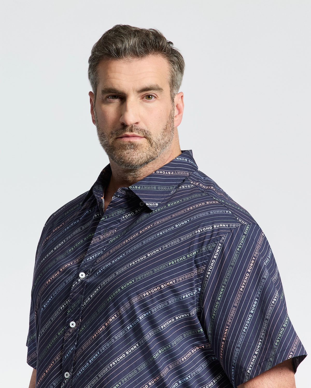 A short-haired, bearded man wears a Psycho Bunny navy blue Renmark Stretch Poplin Woven Shirt with diagonal stripes and text. The premium stretch fabric ensures comfort, while the logo-patterned print adds distinction. He gazes at the camera against a plain background.