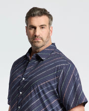 A short-haired, bearded man wears a Psycho Bunny navy blue Renmark Stretch Poplin Woven Shirt with diagonal stripes and text. The premium stretch fabric ensures comfort, while the logo-patterned print adds distinction. He gazes at the camera against a plain background.