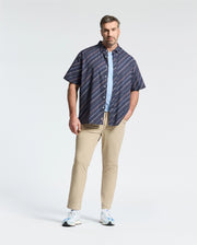 A man in a Psycho Bunny MENS BIG AND TALL RENMARK STRETCH POPLIN WOVEN SHIRT - B9Q629F200, styled over a light blue T-shirt with beige pants and white sneakers, poses with one hand in his pocket against a plain white background.
