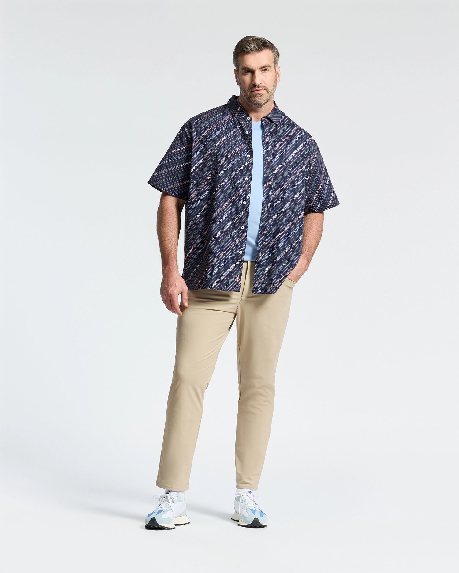 A man in a Psycho Bunny MENS BIG AND TALL RENMARK STRETCH POPLIN WOVEN SHIRT - B9Q629F200, styled over a light blue T-shirt with beige pants and white sneakers, poses with one hand in his pocket against a plain white background.