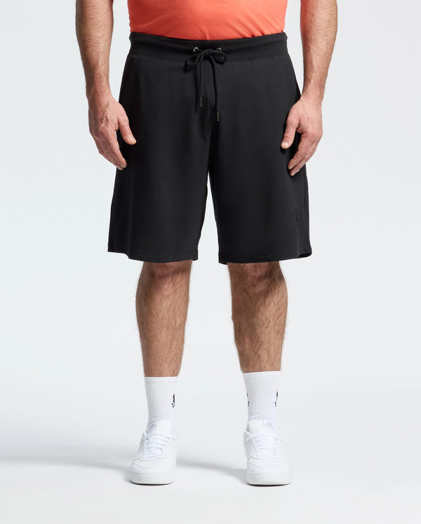 A person stands against a plain white background, wearing a coral shirt, Psycho Bunny MENS BIG AND TALL LEVANZO FRENCH TERRY SWEATSHORT - B9R578F200 in black, white crew socks, and white sneakers. The image is cropped from the shoulders down.