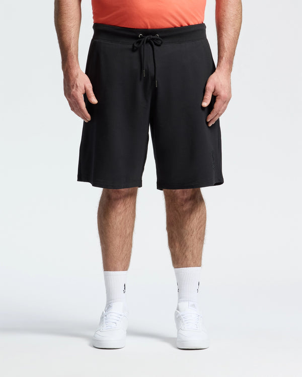 A person stands against a plain white background, wearing a coral shirt, Psycho Bunny MENS BIG AND TALL LEVANZO FRENCH TERRY SWEATSHORT - B9R578F200 in black, white crew socks, and white sneakers. The image is cropped from the shoulders down.