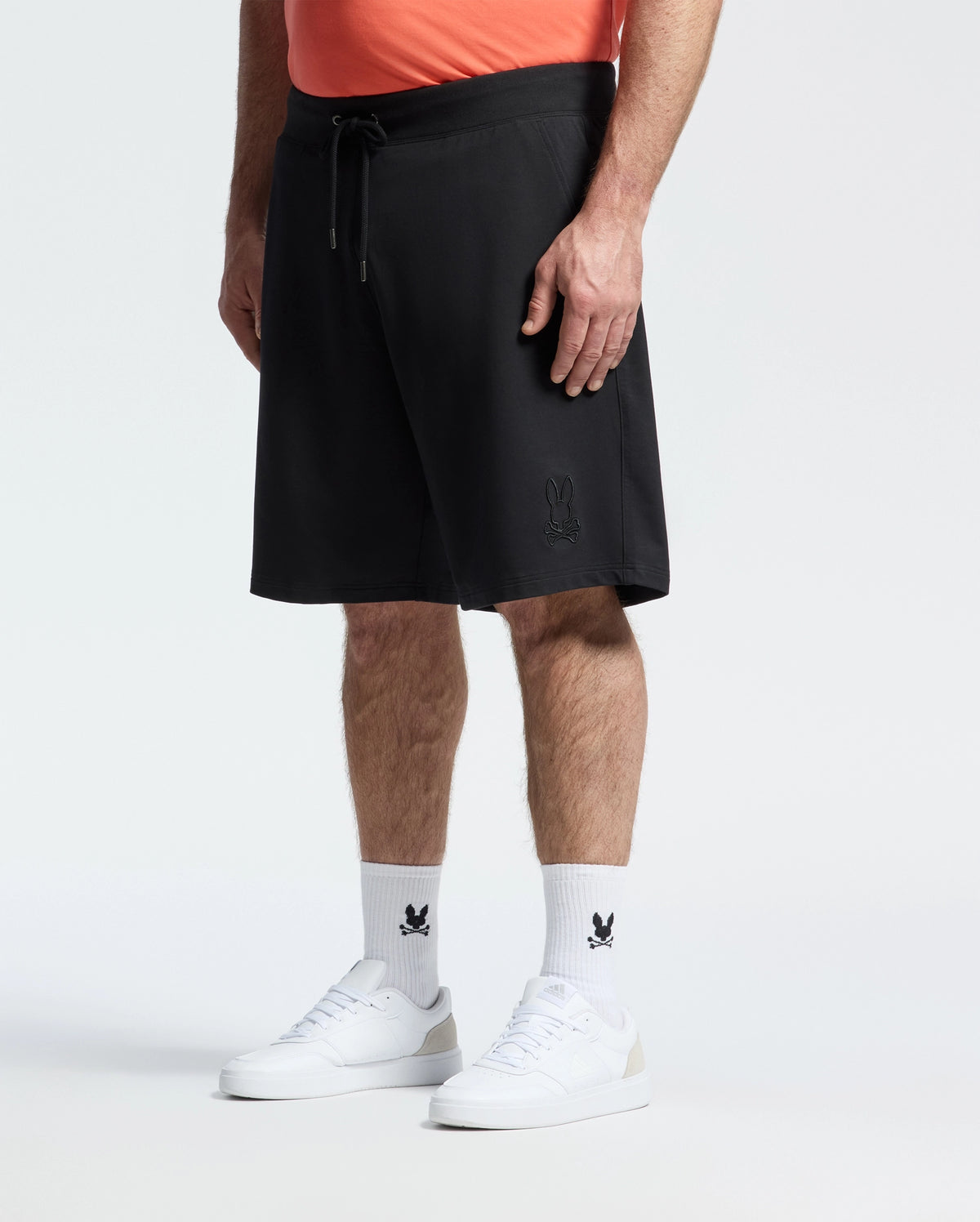 A person stands against a plain light background wearing a coral shirt, Psycho Bunny Mens Big and Tall Levanzo French Terry Sweatshort in black with a small bunny logo, white socks with a black bunny logo, and white sneakers.