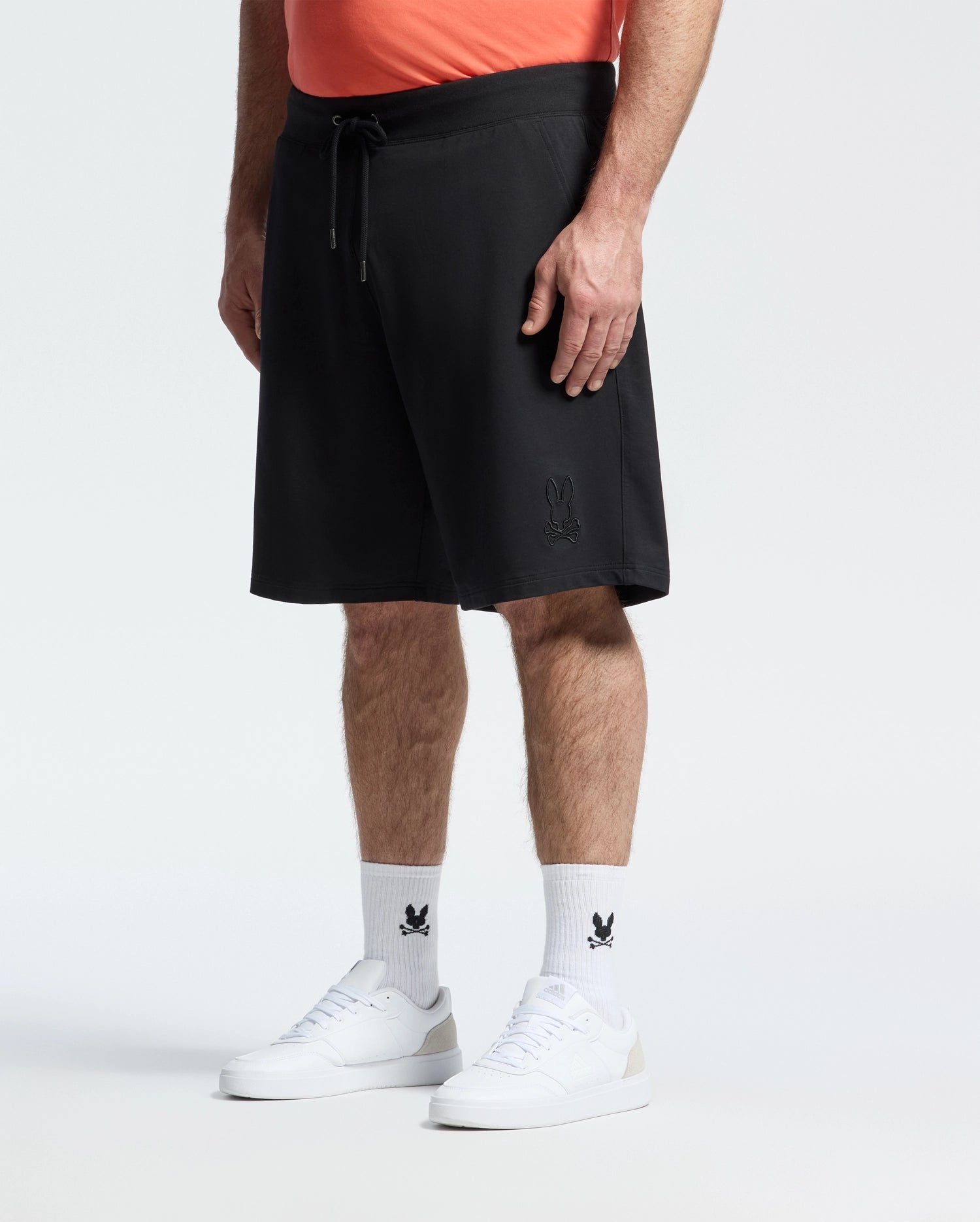 A person stands against a plain light background wearing a coral shirt, Psycho Bunny Mens Big and Tall Levanzo French Terry Sweatshort in black with a small bunny logo, white socks with a black bunny logo, and white sneakers.