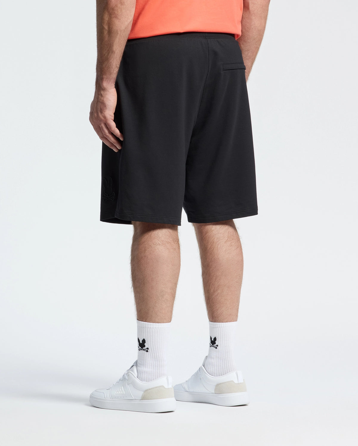 A person wears a coral shirt, white logo socks, white sneakers, and Psycho Bunny MENS BIG AND TALL LEVANZO FRENCH TERRY SWEATSHORT - B9R578F200 in black, standing with their back to the camera against a plain white background.