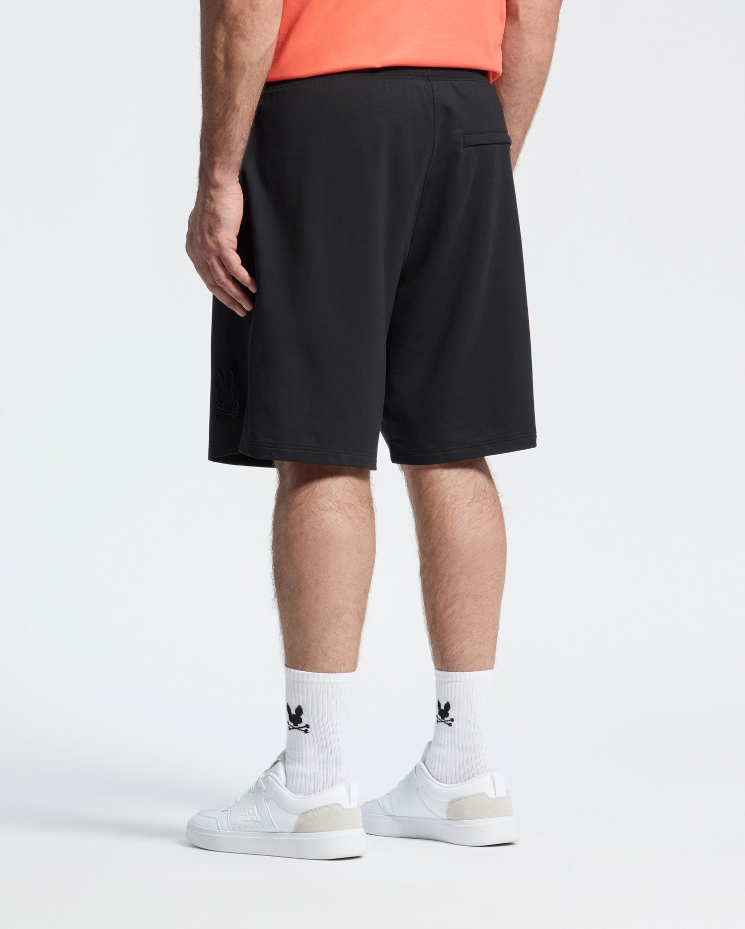 A person wears a coral shirt, white logo socks, white sneakers, and Psycho Bunny MENS BIG AND TALL LEVANZO FRENCH TERRY SWEATSHORT - B9R578F200 in black, standing with their back to the camera against a plain white background.