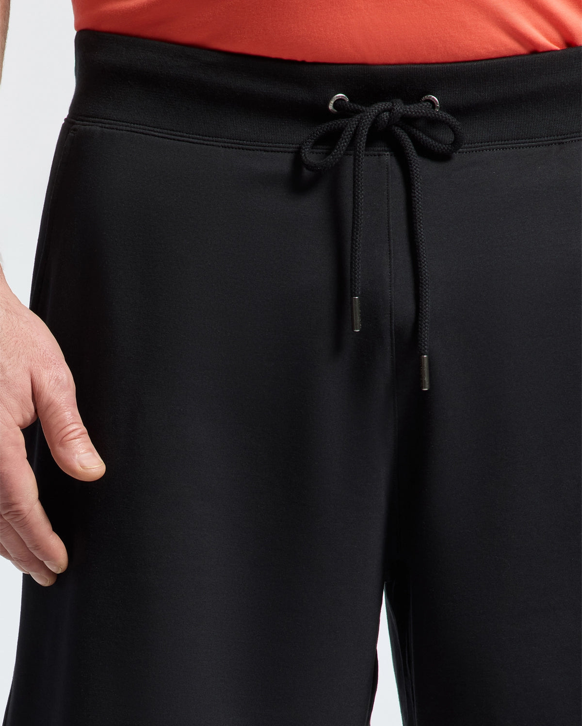 A person wearing Psycho Bunny MENS BIG AND TALL LEVANZO FRENCH TERRY SWEATSHORT - B9R578F200 in black, with only their lower torso and left hand visible against a plain background.