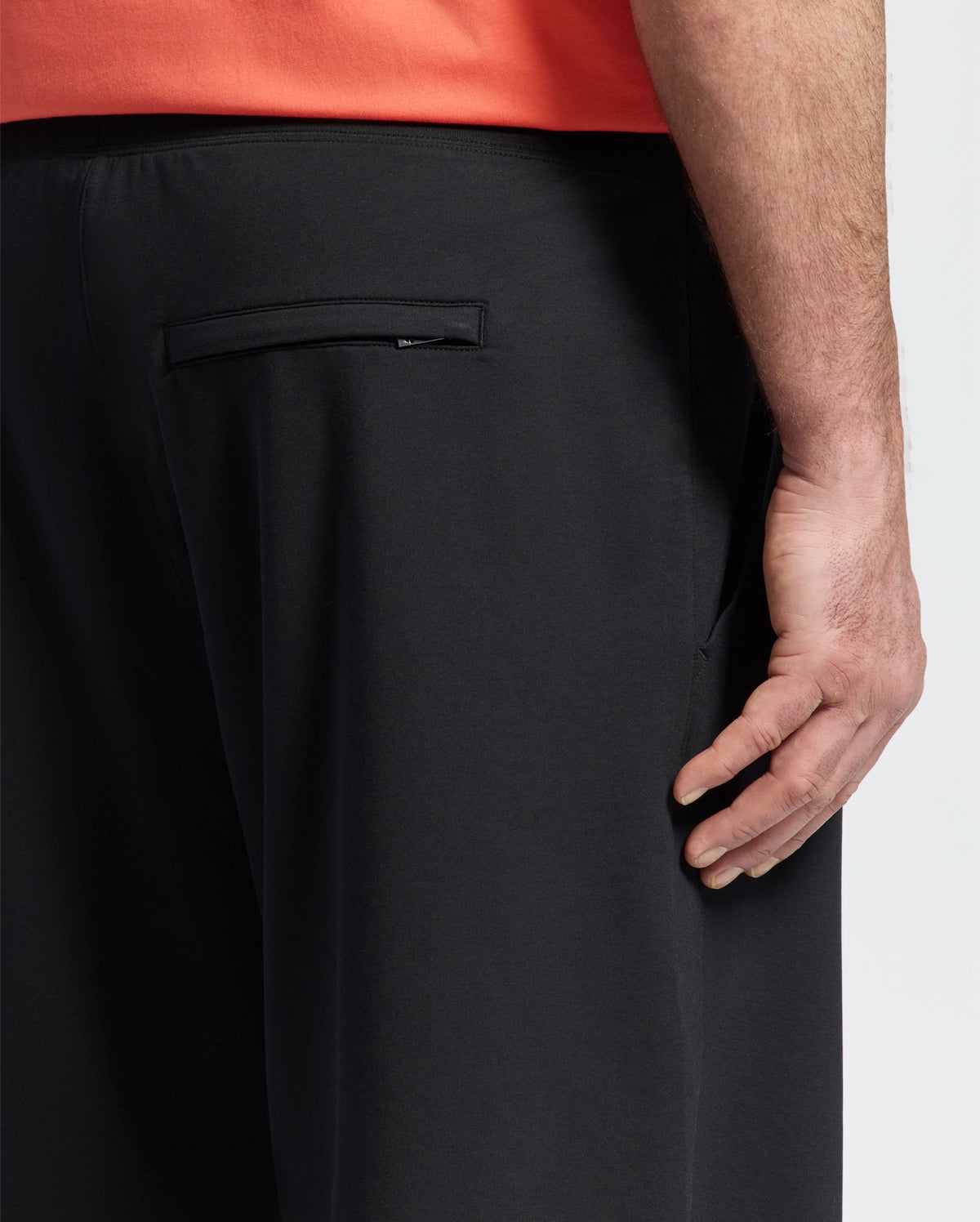 A close-up of a person’s right hand resting on the back pocket of Psycho Bunny MENS BIG AND TALL LEVANZO FRENCH TERRY SWEATSHORT - B9R578F200 in black, with part of an orange shirt visible.