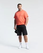A man stands against a light background wearing a coral graphic tee and Psycho Bunny MENS BIG AND TALL LEVANZO FRENCH TERRY SWEATSHORTS (B9R578F200) in black, paired with white socks, white sneakers, and holding a black cap.