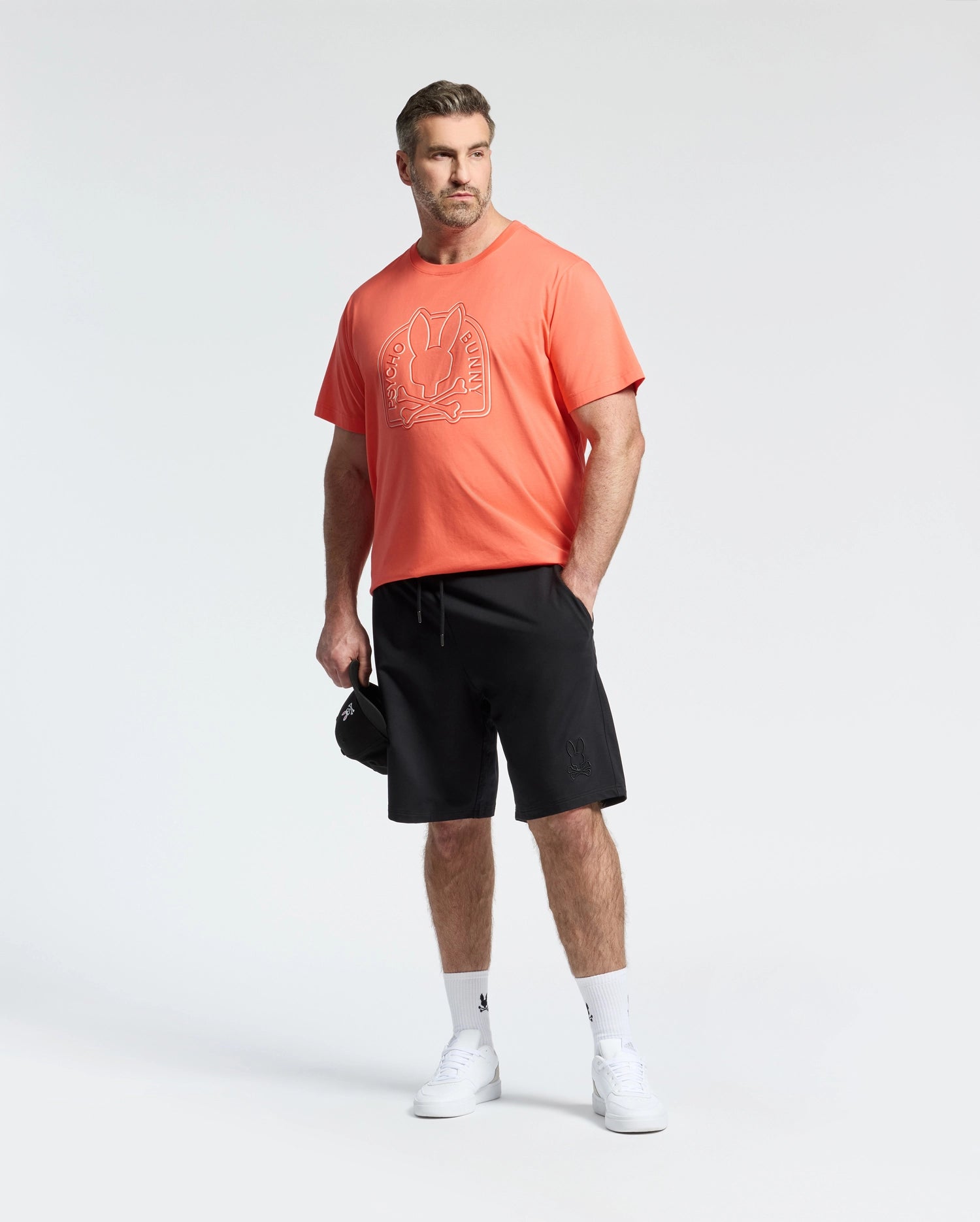 A man stands against a light background wearing a coral graphic tee and Psycho Bunny MENS BIG AND TALL LEVANZO FRENCH TERRY SWEATSHORTS (B9R578F200) in black, paired with white socks, white sneakers, and holding a black cap.