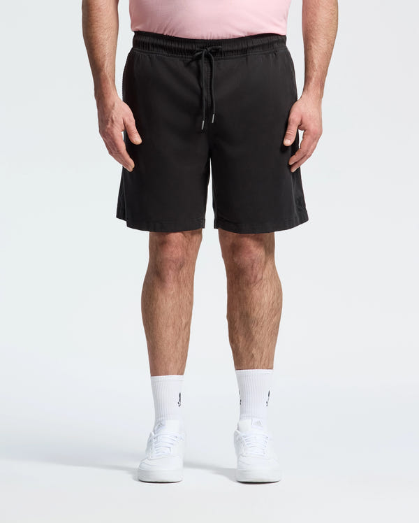A man wears a light pink shirt and black Psycho Bunny Big and Tall Willis Stretch Tencel Shorts (B9R584F200), paired with white crew socks and sneakers. He stands against a plain white backdrop.
