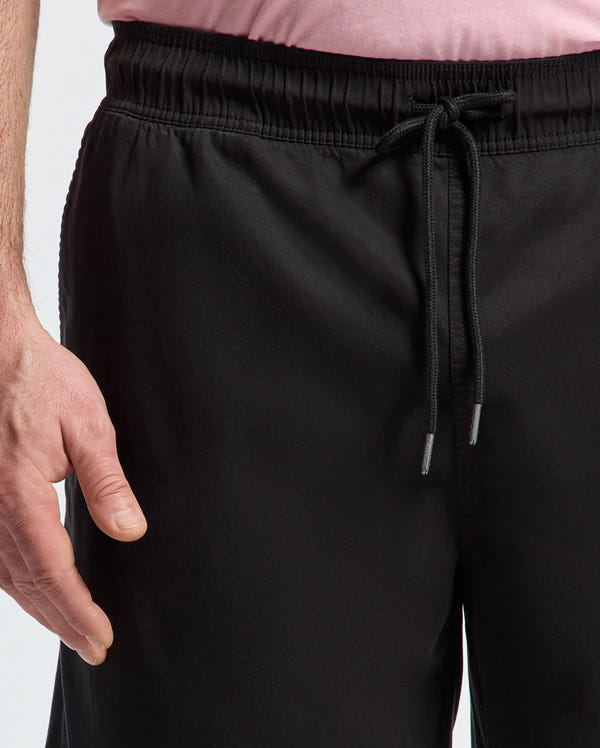 Wearing a light pink shirt and Psycho Bunny black jogger pants with a drawstring, featuring a subtle bunny pattern, the individual has their left hand resting beside the pants, highlighting the waist area of this regular-fit ensemble.