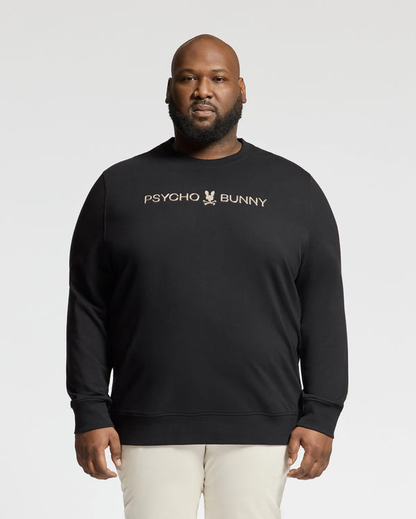 Someone in a black Psycho Bunny Big and Tall Lavata French Terry Sweatshirt (B9S159E200), featuring the logo and text on the front, stands against a plain background, stylishly paired with light-colored pants.