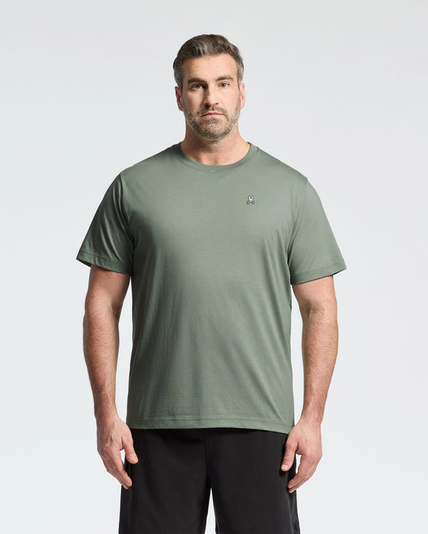 A person with short hair and a beard is wearing Psycho Bunny’s BIG AND TALL CLASSIC CREW NECK TEE in light green with a chest logo, paired with black pants, effortlessly complementing the light gray background.