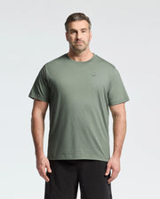 A person with short hair and a beard is wearing Psycho Bunny’s BIG AND TALL CLASSIC CREW NECK TEE in light green with a chest logo, paired with black pants, effortlessly complementing the light gray background.
