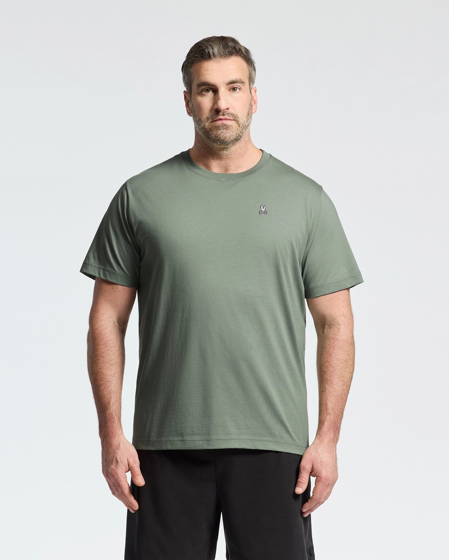 A person with short hair and a beard is wearing Psycho Bunny’s BIG AND TALL CLASSIC CREW NECK TEE in light green with a chest logo, paired with black pants, effortlessly complementing the light gray background.