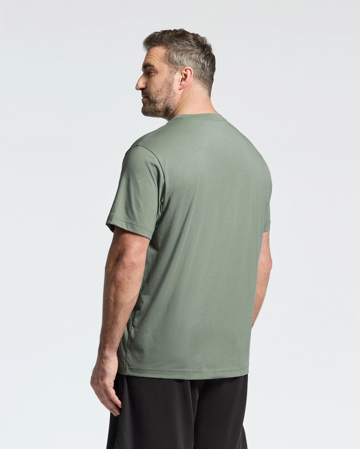 A bearded, short-haired man stands looking left, wearing a Psycho Bunny BIG AND TALL CLASSIC CREW NECK TEE - B9U014CRPC in grayish-green Pima cotton with black pants against a plain white background.