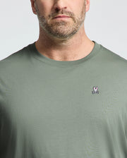 A man with a short beard wears a green BIG AND TALL CLASSIC CREW NECK TEE by Psycho Bunny, displaying a small pink and white bunny logo on the chest, standing out against the light, plain background.