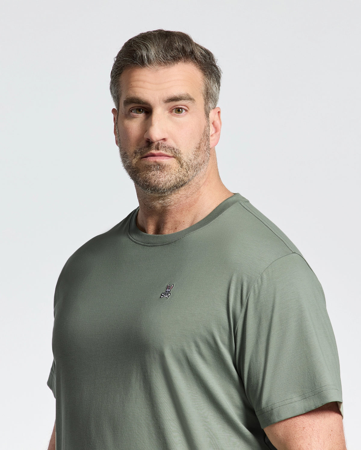 A bearded man with short hair wears a green Psycho Bunny Big and Tall Classic Crew Neck Tee (B9U014CRPC) made from premium Pima cotton, featuring a small embroidered chest logo. He gazes slightly to the side against a plain white background.