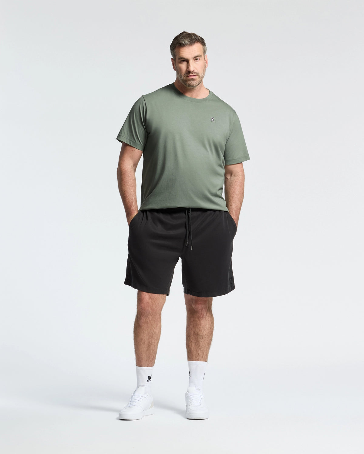 A man stands relaxed against a light background, wearing a soft Pima cotton BIG AND TALL CLASSIC CREW NECK TEE - B9U014CRPC by Psycho Bunny, paired with black shorts and white socks and sneakers. His hands are in his pockets.
