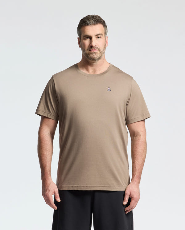 A man with short brown hair and a beard wears the Psycho Bunny BIG AND TALL CLASSIC CREW NECK TEE (B9U014CRPC), featuring a small embroidered logo on the left chest, paired with black shorts against a white background.