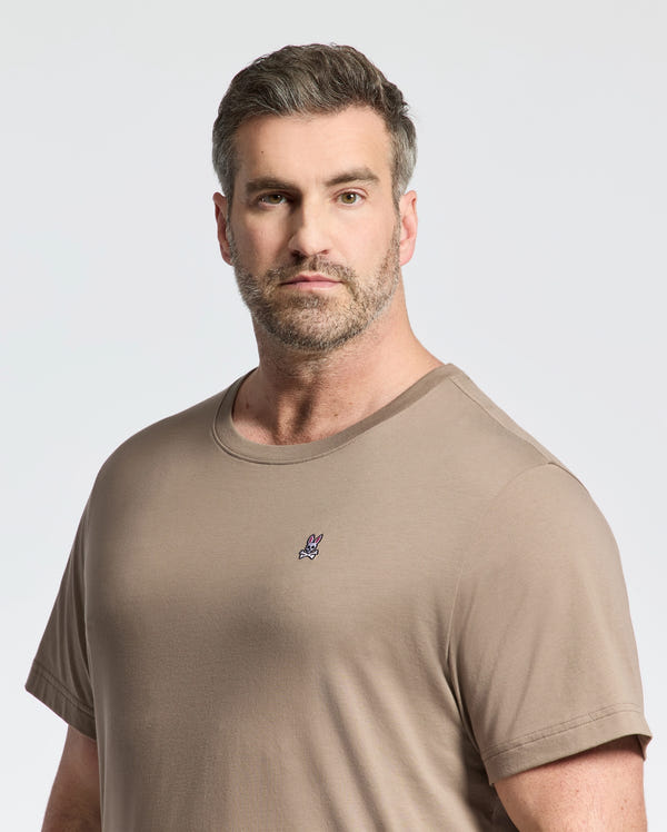 A man with short hair and a beard wears the Psycho Bunny BIG AND TALL CLASSIC CREW NECK TEE (B9U014CRPC) in taupe, made from soft pima cotton and featuring a small rabbit logo on the chest, standing against a plain light background.