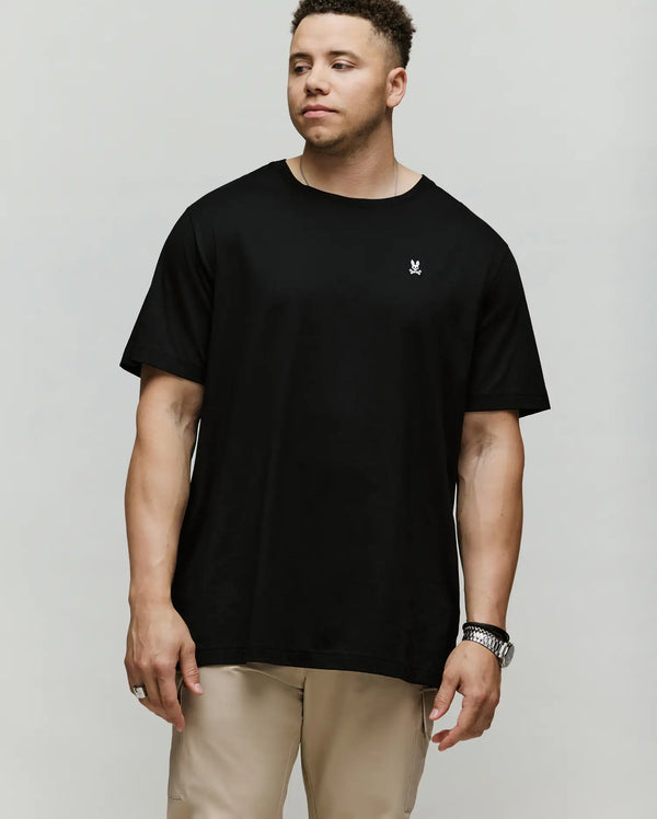 A man with short curly hair wears the Psycho Bunny MENS BIG AND TALL CLASSIC CREW NECK TEE (B9U014CRPC), a loose-fit black 100% Pima cotton shirt with a small white chest logo, and beige pants, standing against a plain light background.