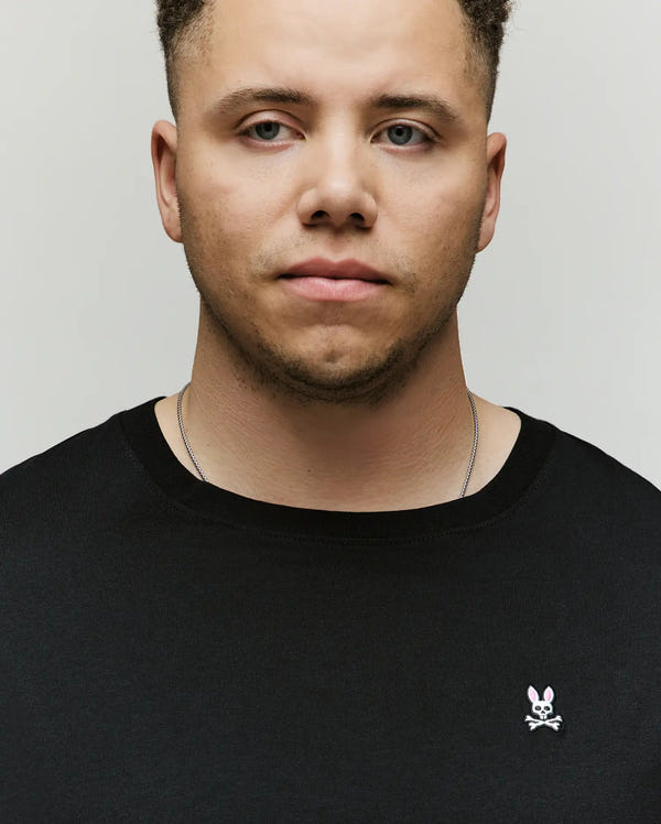 A light-skinned man with short curly hair wears the Psycho Bunny MENS BIG AND TALL CLASSIC CREW NECK TEE (B9U014CRPC) in black, featuring a pink and white bunny skull logo, made from 100% Pima cotton, plus a thin silver necklace against a plain background.