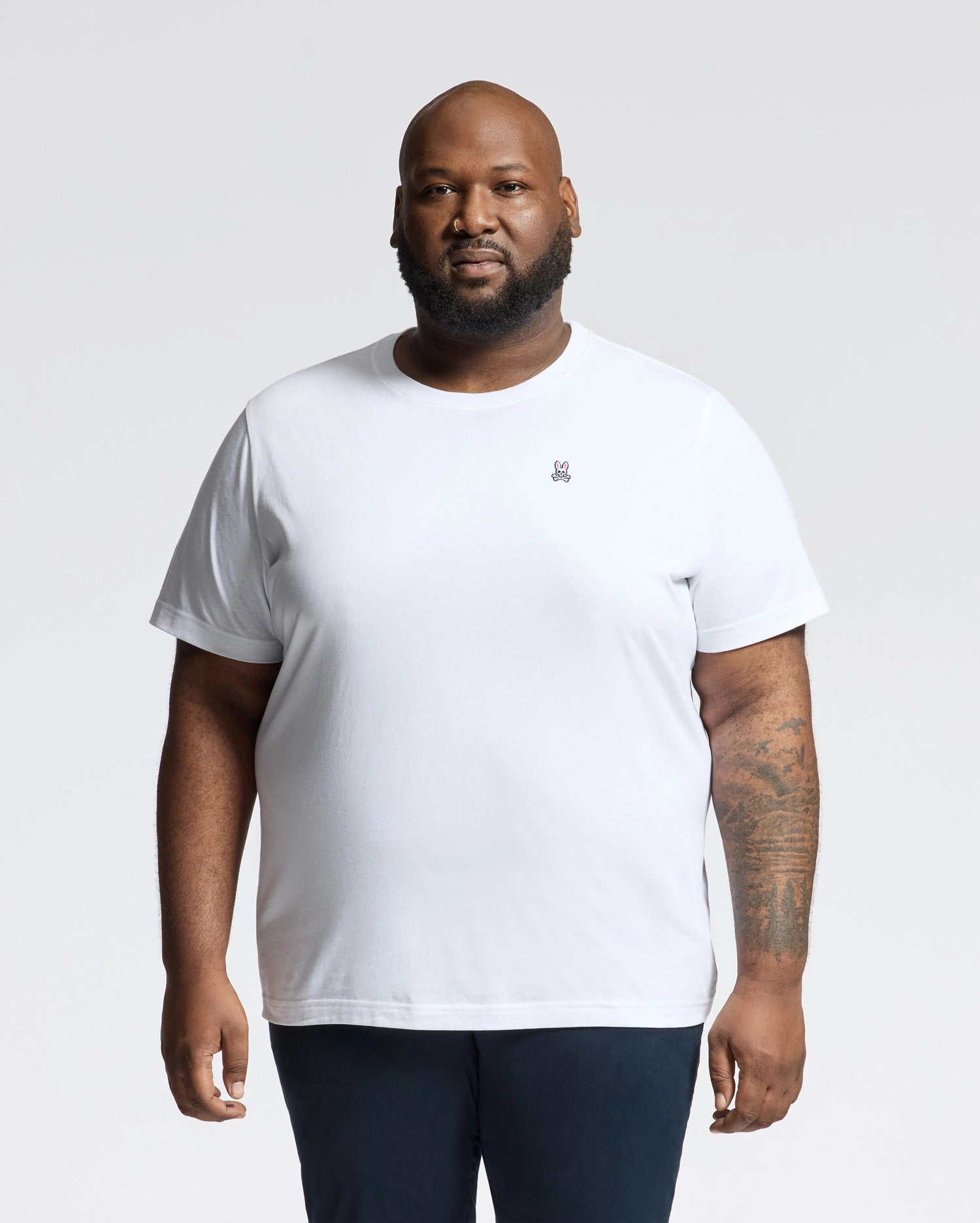 A bearded person with a tattoo on their left forearm wears Psycho Bunny's Men's Big and Tall Classic Crew Neck Tee (B9U014CRPC) in plain white Pima cotton, paired with dark pants, standing against a light gray background, showcasing men's wardrobe essentials.