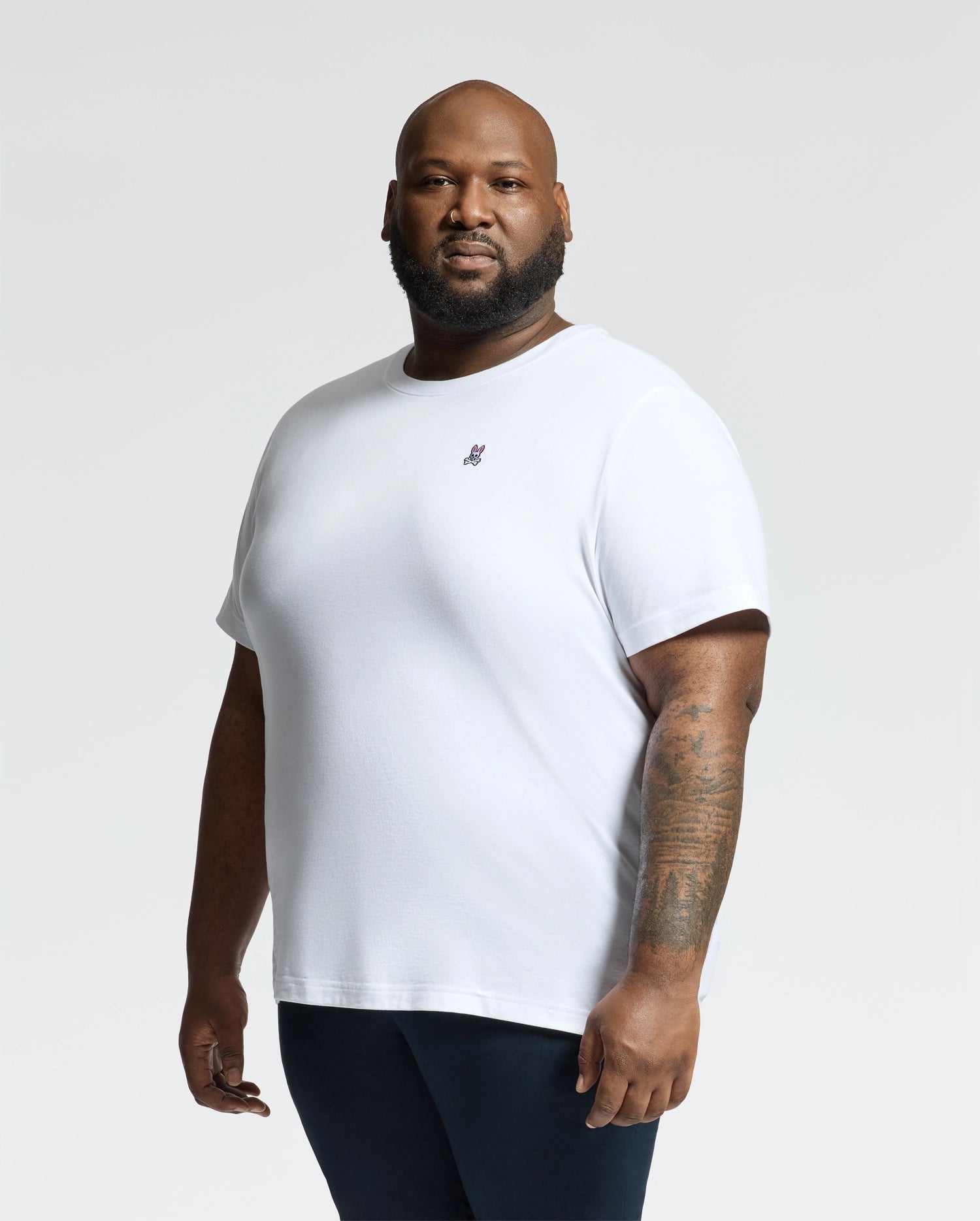 A bearded man showcases the Psycho Bunny MENS BIG AND TALL CLASSIC CREW NECK TEE - B9U014CRPC against a plain background, made from soft Pima cotton with a small embroidered logo, perfectly complementing his dark pants and tattooed arm—essentials in every man's wardrobe.
