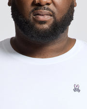 A bearded individual with a nose ring wears the Psycho Bunny MENS BIG AND TALL CLASSIC CREW NECK TEE - B9U014CRPC. It's a white Pima cotton t-shirt with a small pink bunny logo on the chest, set against a neutral backdrop—a timeless addition to any men's wardrobe.