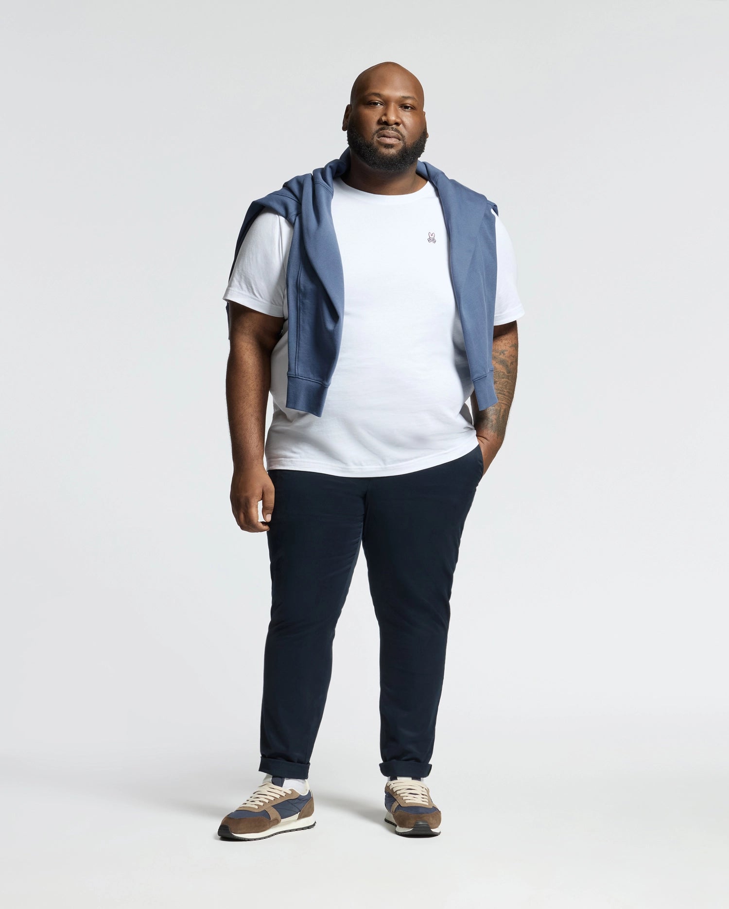 Against a plain backdrop, a person wears a Psycho Bunny MENS BIG AND TALL CLASSIC CREW NECK TEE in white Pima cotton, dark pants, and sneakers. A blue hoodie is draped over their shoulders. Their beard and relaxed posture highlight the versatility of a modern men's wardrobe.
