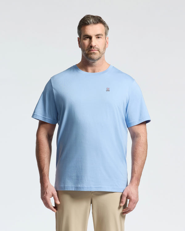 A bearded man with short hair wears a light blue Psycho Bunny BIG AND TALL CLASSIC CREW NECK TEE (B9U014Z1PC) made of 100% Pima cotton, featuring a small embroidered logo on the left chest. He faces forward against a plain light background, paired with beige pants for a timeless look.
