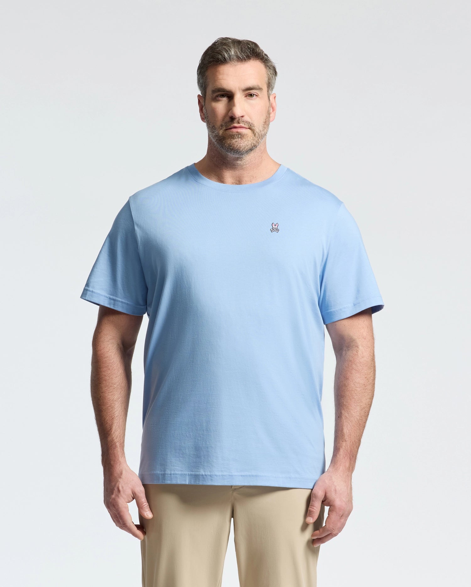 A bearded man with short hair wears a light blue Psycho Bunny BIG AND TALL CLASSIC CREW NECK TEE (B9U014Z1PC) made of 100% Pima cotton, featuring a small embroidered logo on the left chest. He faces forward against a plain light background, paired with beige pants for a timeless look.