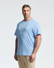 A man with short brown hair and facial hair stands against a light background wearing Psycho Bunny's Big and Tall Classic Crew Neck Tee in light blue, made from 100% Pima cotton for ultimate comfort, paired with beige pants. His expression is neutral.