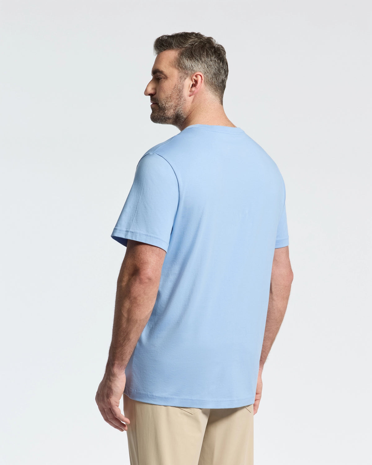 A bearded man with short hair stands slightly to the right, wearing a Psycho Bunny BIG AND TALL CLASSIC CREW NECK TEE (B9U014Z1PC) in light blue, made of 100% Pima cotton, paired with beige pants. The background is plain white.