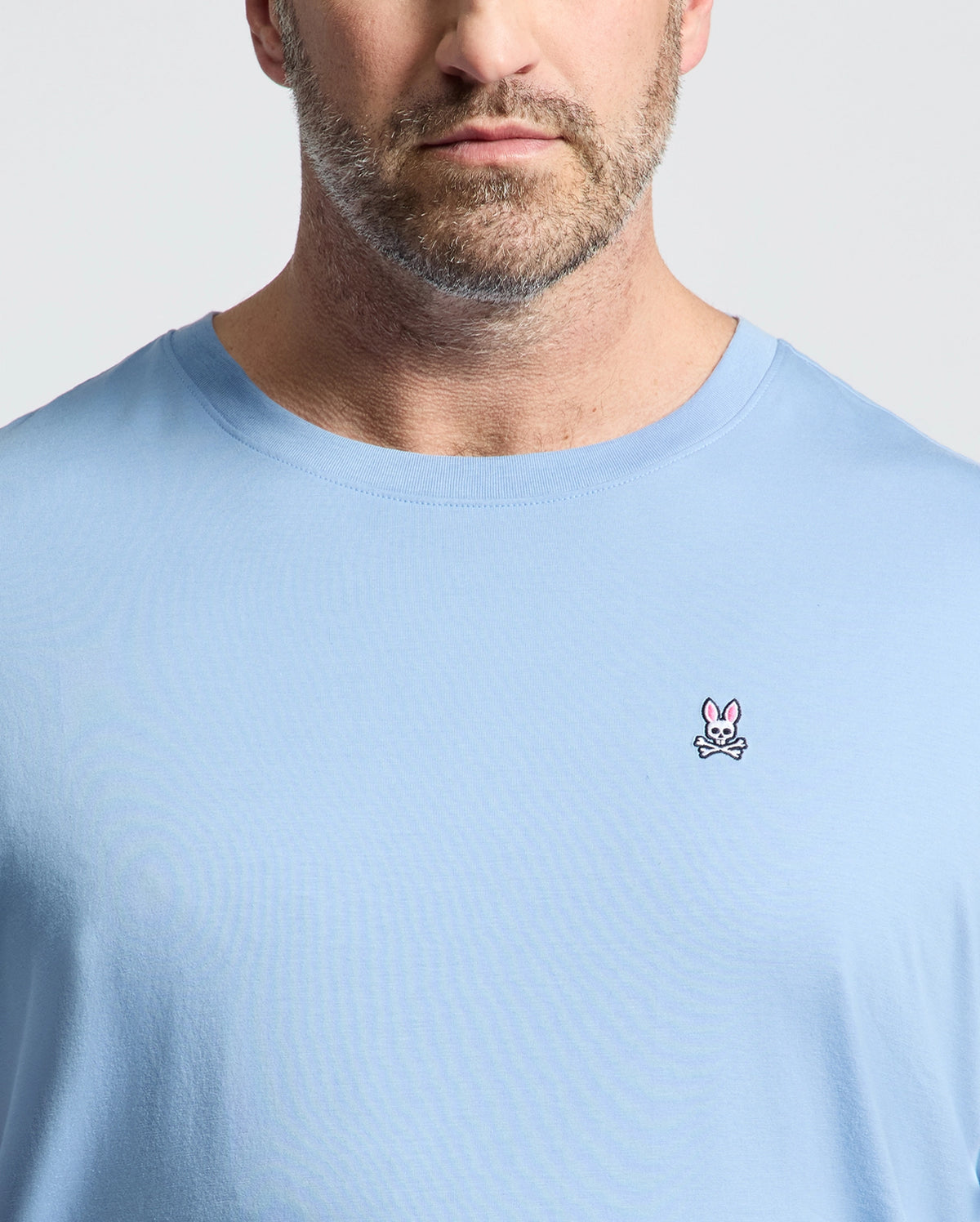 A man wears Psycho Bunny's BIG AND TALL CLASSIC CREW NECK TEE (B9U014Z1PC) made of 100% Pima cotton, in light blue, featuring a pink rabbit and crossed bones logo on the left chest, against a plain light background.