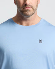 A man wears Psycho Bunny's BIG AND TALL CLASSIC CREW NECK TEE (B9U014Z1PC) made of 100% Pima cotton, in light blue, featuring a pink rabbit and crossed bones logo on the left chest, against a plain light background.