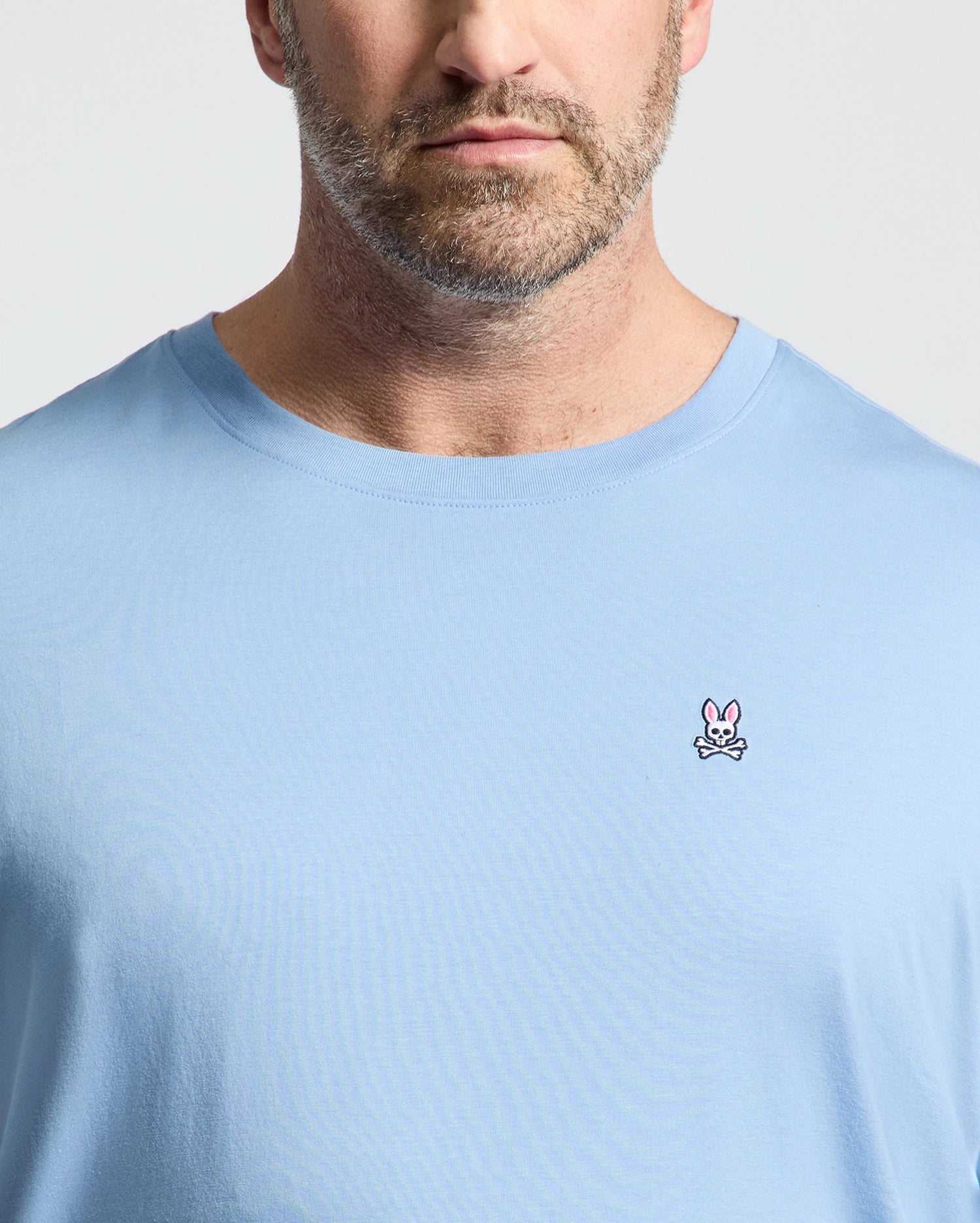 A man wears Psycho Bunny's BIG AND TALL CLASSIC CREW NECK TEE (B9U014Z1PC) made of 100% Pima cotton, in light blue, featuring a pink rabbit and crossed bones logo on the left chest, against a plain light background.